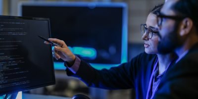 diverse AI developers collaborate in a late-night coding session. A senior engineer mentors his colleague as they program a complex machine learning algorithm together.
